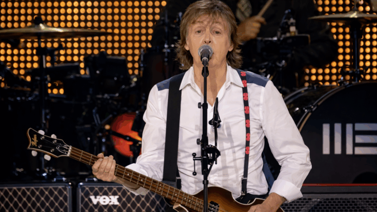 Paul McCartney performs on stage, singing into a microphone while playing his iconic bass guitar against a bright backdrop of concert lights.