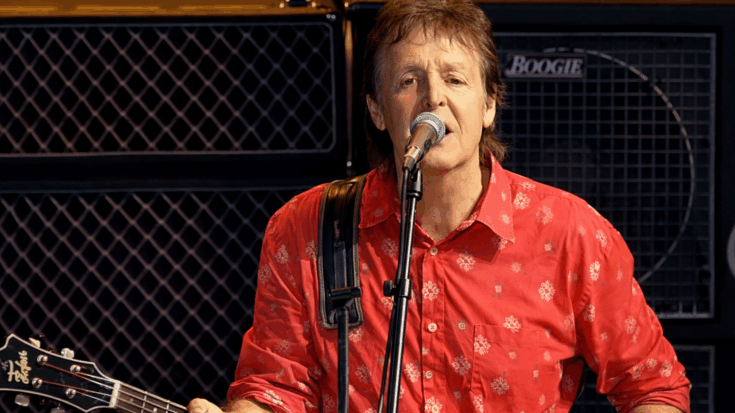 Paul McCartney sings into a microphone while playing his bass guitar on stage, wearing a bright red shirt.