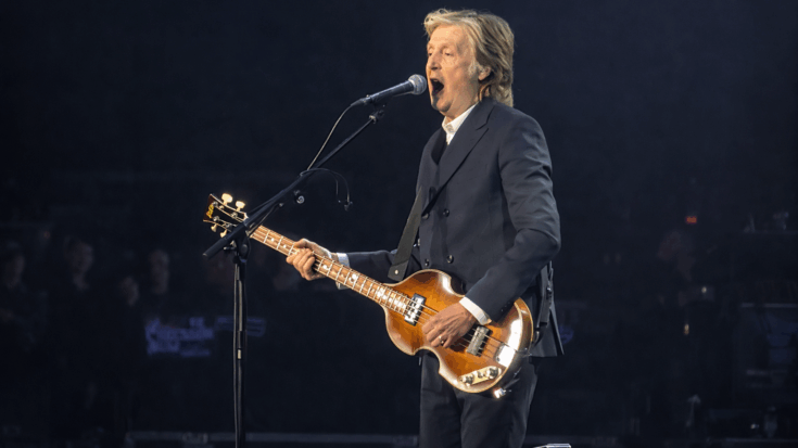 Paul McCartney sings into a microphone while playing his bass guitar on stage under soft concert lighting.