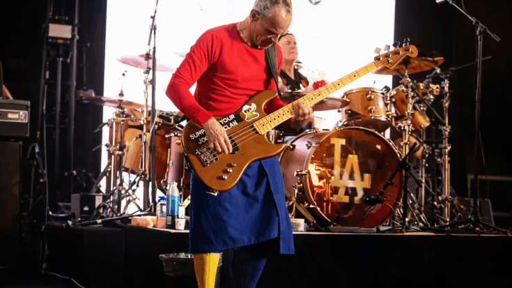 Flea is an energetic bassist performing onstage in a bright red shirt, colorful leggings, and a sticker-covered bass.