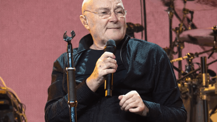 Phil Collins holds a microphone on stage, looking to the side with a calm expression against a warm concert backdrop.