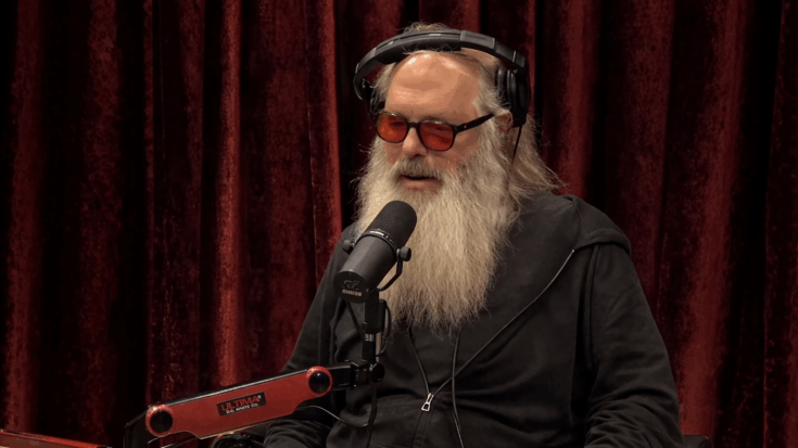 Rick Rubin sits in a podcast studio wearing headphones and tinted glasses, speaking into a microphone with his long white beard flowing over a dark hoodie.