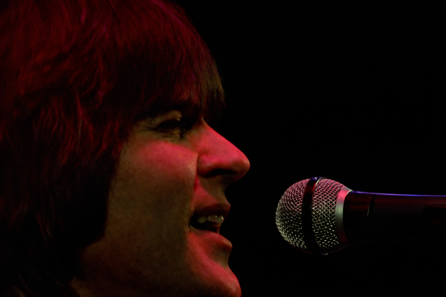 Led Randy is shown in a close-up side profile, singing into a microphone under dark red stage lighting.