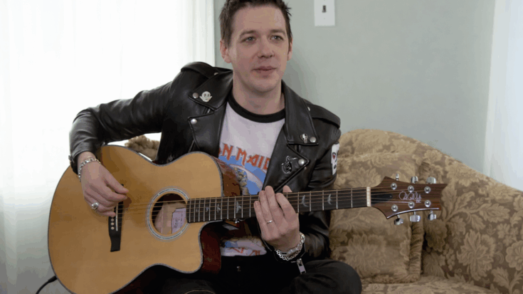 Tobias Forge sits casually on a patterned couch, holding an acoustic guitar while wearing a black leather jacket and graphic shirt, appearing relaxed as if mid-conversation or demonstration.
