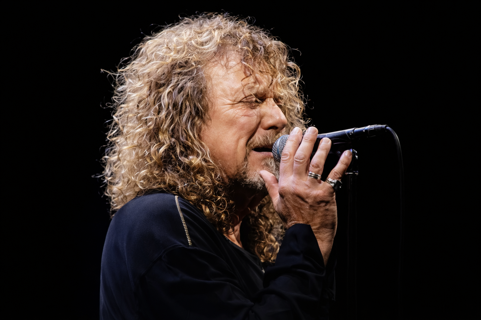 Robert Plant sings passionately into a microphone, his curly hair framing his face as he performs on a dark stage.