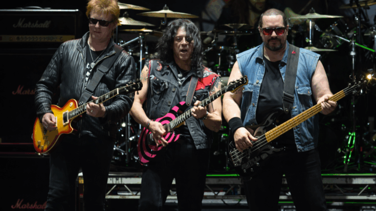 Twisted Sister performs onstage with three members playing guitars and bass under bright concert lights.