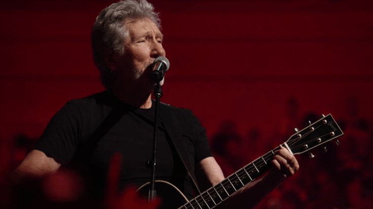 Roger Waters appears focused and intense, performing under moody blue lighting with a serious stage presence.