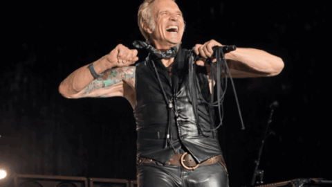 David Lee Roth beams onstage, flexing his arms while holding a microphone mid-performance.
