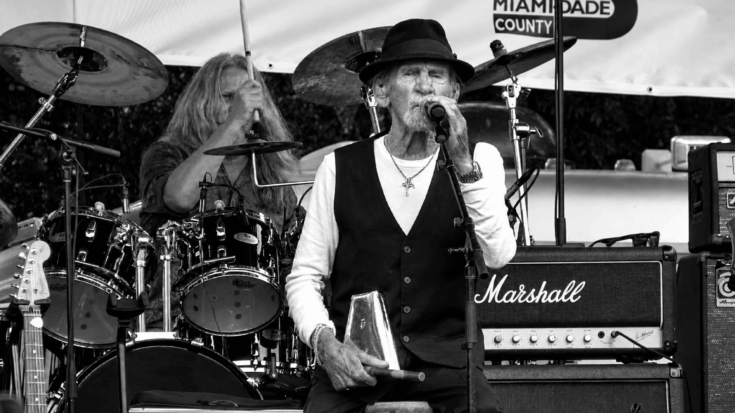 Alex Ligertwood performs on stage in black and white, singing into a microphone while holding a percussion instrument as the band plays behind him.